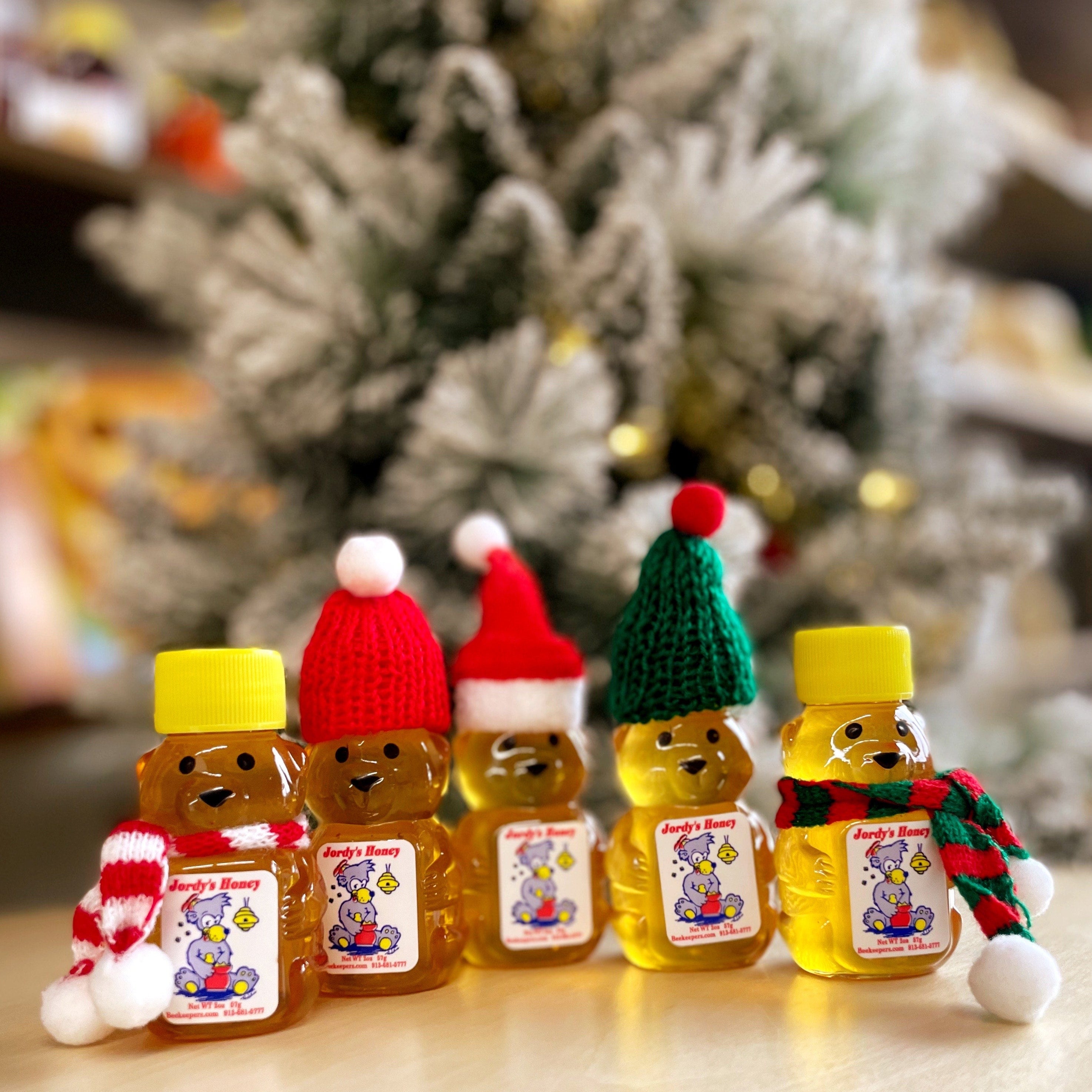 Five bear-shaped honey jars with knit hats and scarves in front of a decorated Christmas tree.